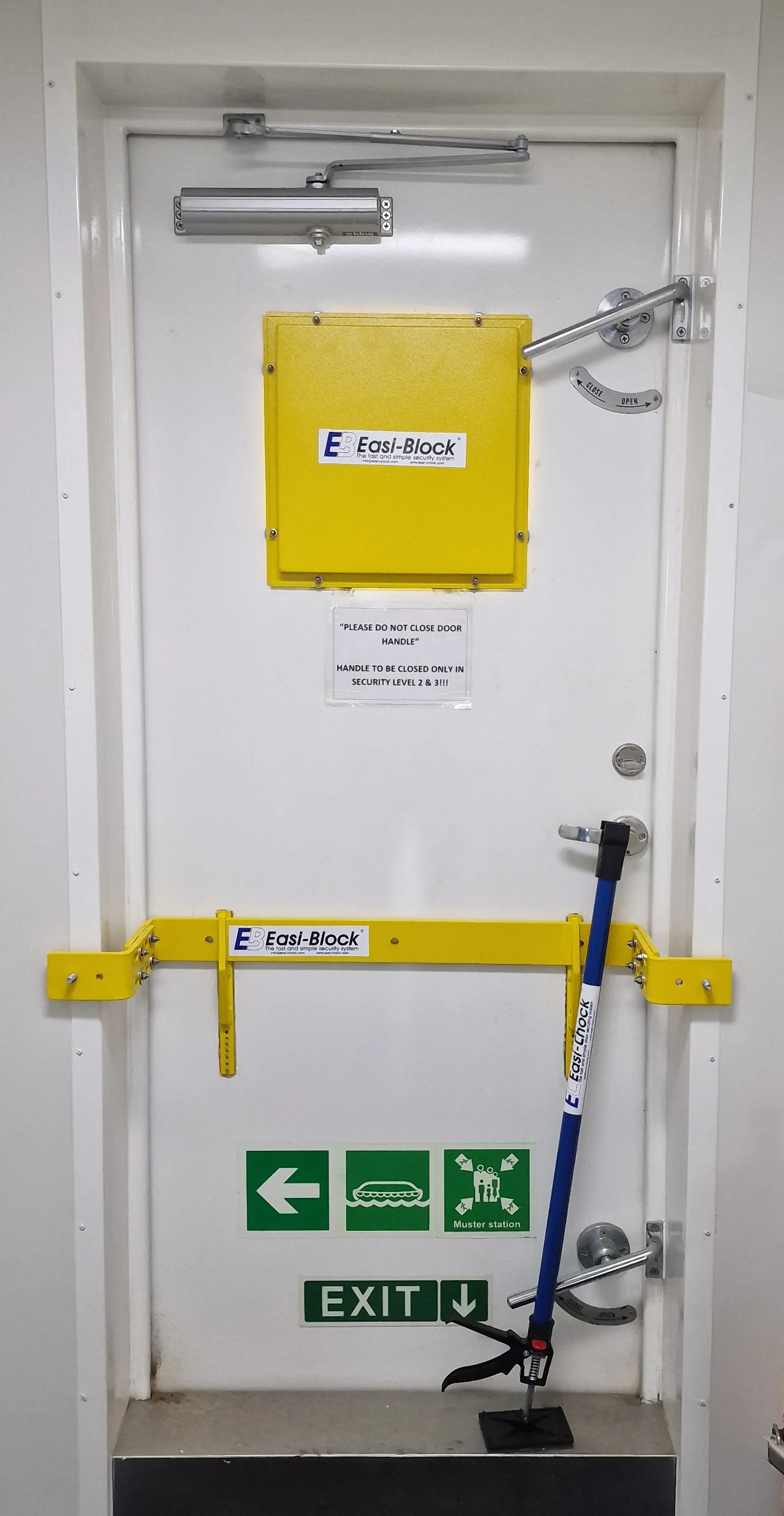 A steel ship's door by a muster station, its window plated over and held shut by a yellow Easi-Block bar and a blue Easi-Chock